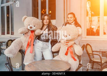 L'amicizia delle donne orsi tiene in mano giganteschi giocattoli di peluche in una terrazza soleggiata mentre sorridono insieme durante l'ora d'oro Foto Stock