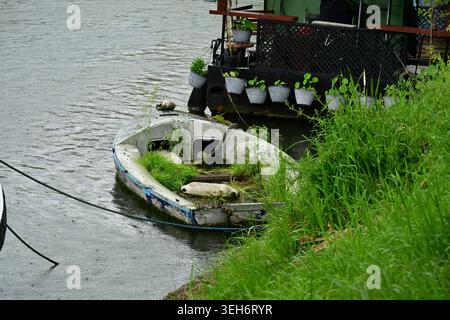 Barca sul fiume abbandonata ricoperta di erba accanto al molo galleggiante del giardino Foto Stock