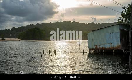 La luce del sole dorata irrompe attraverso nuvole di tempesta sopra una baia vietnamita, scintillando sull'acqua accanto a un palafitte ondulato arrugginito con vecchi pali di legno. Vie Foto Stock