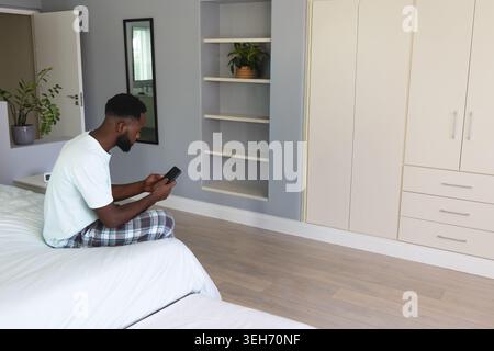 Uomo afroamericano medio adulto seduto sul letto tenendo il telefono, guardando lo schermo in camera, fotocopiatrice Foto Stock