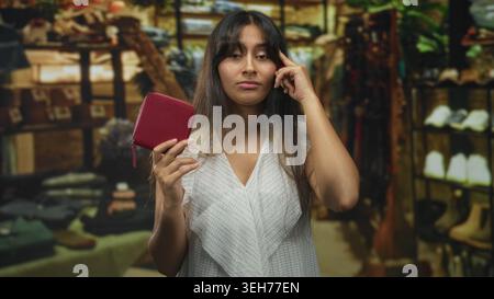 Bruna latina giovane donna che tiene un piccolo portafoglio rosso, dito al tempio mentre esamina gli scaffali in un negozio di edifici; contemplazione tranquilla. Foto Stock