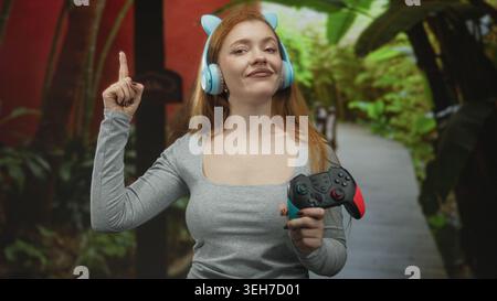 Giovane donna rossa che tiene in mano il gamepad wireless e indossa le cuffie blu Cat, punta il dito indice nella foresta; giocosa. Foto Stock