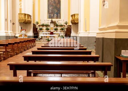 Sessa Aurunca - Campania - Italia - 08-29-2025: File di pali di legno vuoti conducono verso un altare ornato con pulpiti dorati e piante all'interno di una luminosa chiesa storica di Sessa Aurunca. Foto Stock