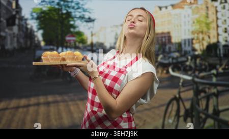 Una donna bionda che indossa un grembiule di Vichy rosso che tiene con le mani un vassoio di muffin in legno e sorride per strada; ospitalità gioiosa. Foto Stock
