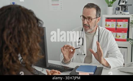 Uomo di mezza età medico con stetoscopio tiene la penna e i gesti mentre consulta il paziente alla scrivania con computer e cartella medica nell'edificio della clinica; re Foto Stock