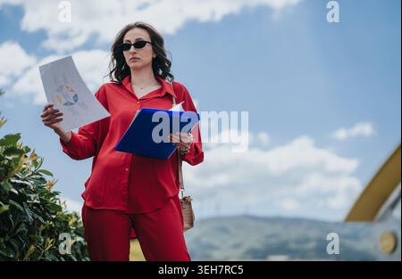 Donna d'affari in abito rosso che esamina documenti all'aperto con cartella blu e occhiali da sole Foto Stock