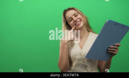 Donna bionda giovane che tiene gli appunti e punta il dito verso l'alto mentre sorride in studio; curiosità giocosa. Foto Stock