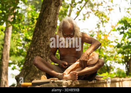 Sri Lanka - 12 febbraio 2026: Vista di un uomo anziano con una lunga barba bianca che intaglia una scultura in legno seduto all'aperto sotto lussureggianti alberi verdi. Foto Stock