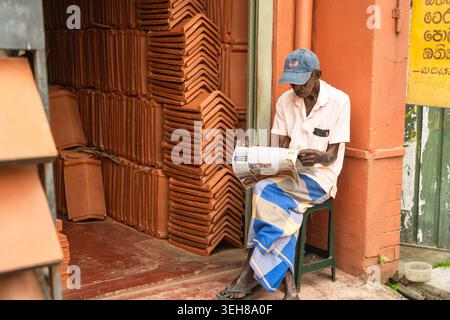 Sri Lanka - 12 febbraio 2026: Veduta di un anziano che legge un giornale seduto su uno sgabello accanto a pile di tegole di terracotta all'esterno di una sh Foto Stock