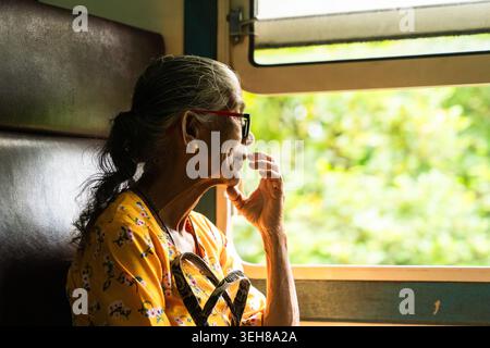 Sri Lanka - 06 febbraio 2026: Veduta di una donna anziana seduta su un treno e che guarda fuori dalla finestra il lussureggiante paesaggio verde. Foto Stock