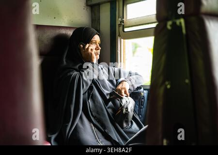 Sri Lanka - 06 febbraio 2026: Vista di una donna in un hijab nero che parla su un telefono cellulare mentre è seduta vicino alla finestra all'interno di un vagone ferroviario. Foto Stock