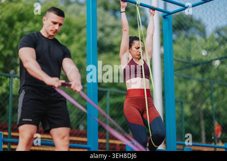 Allenamento fitness: Donna che fa il pull-up assistito con fascia di resistenza mentre l'allenatore tiene le fasce all'aperto Foto Stock
