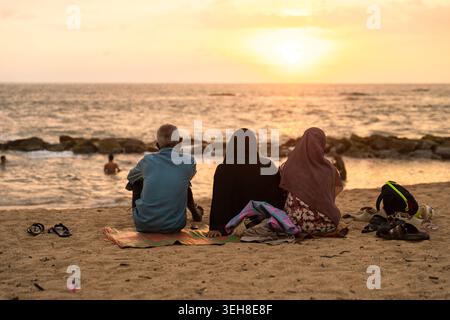 Sri Lanka - 14 gennaio 2026: Veduta di tre persone sedute su una spiaggia sabbiosa che guarda il tramonto sull'Oceano Indiano con frangiflutti rocciosi e nuotate Foto Stock