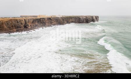 Vista aerea della storica Fortezza di Sagres arroccata su aspre scogliere con potenti onde che si infrangono contro la riva sotto un cielo coperto a Sagres, F. Foto Stock