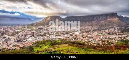 Riprese grandangolari con vista sulla densa architettura urbana di città del Capo ai piedi di Table Mountain, con spettacolari nuvole pesanti e una collina erbosa Foto Stock
