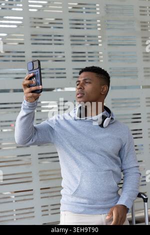 Ragazzo afroamericano che scatta selfie con il telefono a una parete di metallo e indossa una cuffia con cappuccio e custodia Foto Stock
