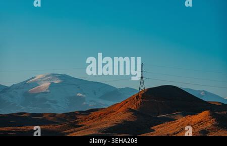 Linee elettriche solitarie sulla collina arancione con montagne innevate al tramonto Foto Stock