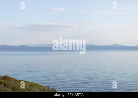 Ampia vista panoramica di un mare blu calmo con sottili increspature, incorniciata da una catena montuosa lontana e un cielo azzurro con nuvole sparse. Foregroun Foto Stock