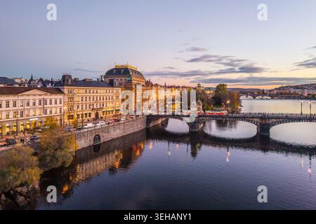 Vista aerea del Teatro Nazionale di Praga durante la serata. L'edificio è illuminato e si riflette nel fiume. Ponti e strade mostrano la città Foto Stock