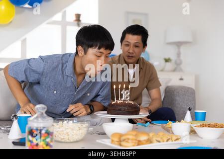 Padre e figlio asiatici festeggiano in salotto, soffiando candele sulla torta al cioccolato con 18 topper Foto Stock