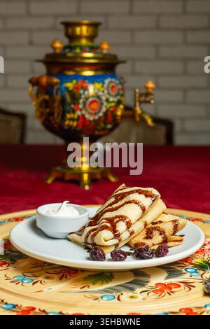 Un piatto di crepes dolci con sciroppo di cioccolato siede su un vassoio decorato di fronte a un samovar tradizionale dipinto Foto Stock