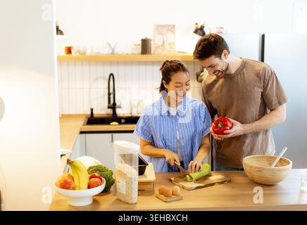 Una donna coreana e un uomo caucasico cucinano in una cucina moderna. Sta tritando verdure mentre lui tiene un pepe rosso. Sembrano felici ed enga Foto Stock