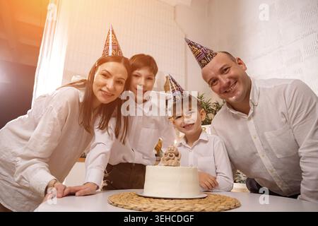 Famiglia sorridente intorno alla torta di compleanno con bambini che indossano cappelli da festa, genitori e bambini riuniti al tavolo, candele accese, cucina che riempie la luce del sole, gioioso Foto Stock