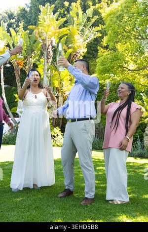 Sposa con gli ospiti del matrimonio che indossano abito e velo alzando flauti e brindando sul prato con champagne. All'aperto, sul retro, tropicale, verde, festeggiamenti, elegante, schietto Foto Stock