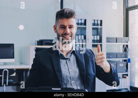 Un uomo d'affari sorridente in tuta lavora su un computer portatile in un ufficio moderno e luminoso Foto Stock
