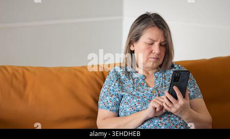 Donne che leggono un messaggio. Donna preoccupata che utilizza il telefono mentre è seduta sul divano Vibrant Orange. La donna moderna appare seria e attenta mentre è impegnata Foto Stock