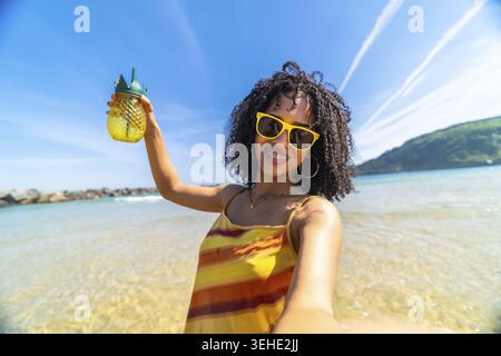 Giovane donna birazziale con bretelle e occhiali da sole che scatta un selfie, tiene in mano una tazza di ananas e sorride brillantemente in una soleggiata giornata estiva in spiaggia, con Foto Stock