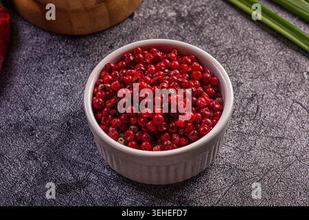Primo piano di grani di pepe rosa rosso brillante in un recipiente di ceramica bianco. Sfondo rustico scuro con peperoncino rosso sfocato, mortaio di legno, cipolla verde Foto Stock