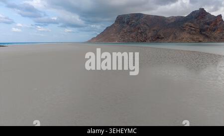 Ampia spiaggia di sabbia vuota con acque poco profonde e un'aspra montagna rocciosa sotto il cielo nuvoloso, calmo paesaggio costiero. Foto Stock