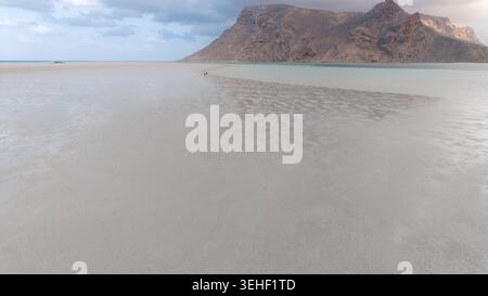 Calmo paesaggio marino con ampie acque poco profonde e sabbia ondulata che conduce a una montagna rocciosa lontana sotto un cielo nuvoloso, con una piccola persona che cammina lontano. Foto Stock