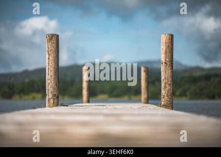 Un molo di legno con quattro pali si estende in un lago tranquillo, con colline verdi sfocate e un cielo nuvoloso che crea un'atmosfera serena Foto Stock
