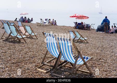 Soleggiata giornata estiva sulla spiaggia di ciottoli di Brighton con file di colorate sedie a sdraio a righe, ombrelloni rossi Coca Cola e persone che si rilassano in riva al mare Foto Stock