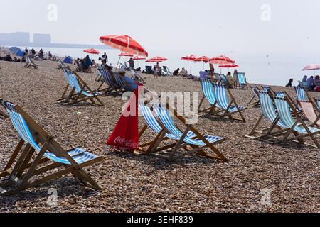 Soleggiata giornata estiva sulla spiaggia di ciottoli di Brighton con file di colorate sedie a sdraio a righe, ombrelloni rossi Coca Cola e persone che si rilassano in riva al mare Foto Stock