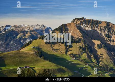 Prato alpino illuminato dal sole punteggiato da chalet e tortuose strade agricole sotto aspre cime innevate. Foto Stock