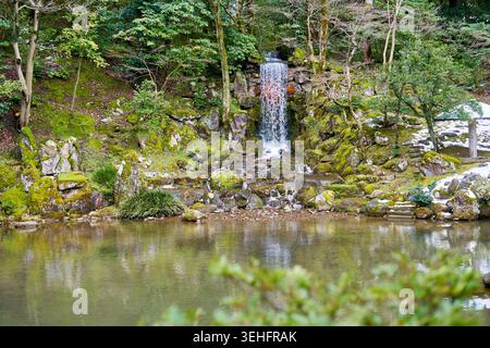 Una scena serena di giardino giapponese con una piccola cascata che scende su rocce ricoperte di muschio in uno stagno calmo. La cascata è circondata da lussureggiante Foto Stock