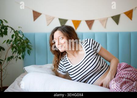 Donna incinta sdraiata sul fianco a letto con un cuscino di maternità e si sente a suo agio, godendo un momento di relax nella sua luminosa camera da letto durante la gravidanza Foto Stock