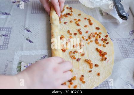Le mani arrotolano l'impasto cosparso di uvetta, un passo avanti nella preparazione del Kozunak, un tradizionale pane dolce. La parola Lavande è visibile sulla tovaglia. Foto Stock