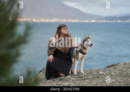 Una donna siede accanto a un cane Husky siberiano sulle rocce costiere. Il mare blu, il paesaggio montano e la postura rilassata creano un'atmosfera di vita tranquilla Foto Stock