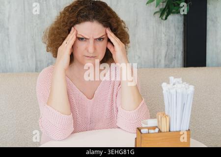 Donna di mezza età con mal di testa ed espressione stressata seduta ad un tavolo da caffè con un maglione rosa Foto Stock