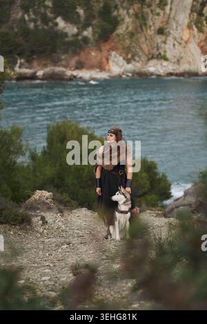 Una donna cammina con un cane Husky siberiano vicino al mare. Il paesaggio naturale con le scogliere e la luce soffusa del giorno crea un'autentica sensazione di stile di viaggio Foto Stock