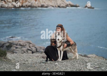 Una donna e un cane Siberian Husky riposano insieme sulle rocce del mare. Il paesaggio costiero con acqua blu e montagne lontane crea un ambiente cinematografico all'aperto Foto Stock