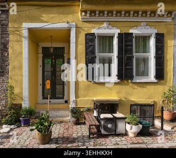Facciata dell'edificio di colore giallo luminoso con decorazioni bianche, porta nera e finestre con persiane. Piante in vaso, pavimentazione a motivi geometrici e un'unità AC completano questa tradizionale vista sulla strada mediterranea Foto Stock