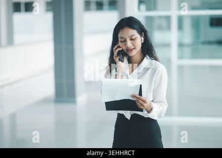 Donna professionista che parla con uno smartphone mentre esamina i documenti in un luminoso corridoio dell'ufficio, mostrando le comunicazioni con i clienti, il multitasking e i documenti Foto Stock