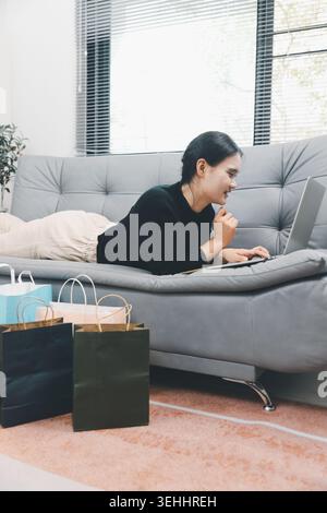 Giovane donna asiatica sdraiata sul divano che usa il laptop per fare shopping online a casa con borse colorate, sfogliare offerte di e-commerce e fare shopping al dettaglio digitale Foto Stock
