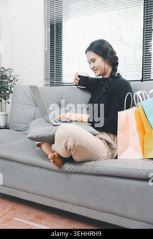 Giovane donna asiatica che usa un computer portatile e beve un caffè sul divano a casa, lavora da remoto e concetto di stile di vita online, ufficio domestico informale, prodotti freelance Foto Stock