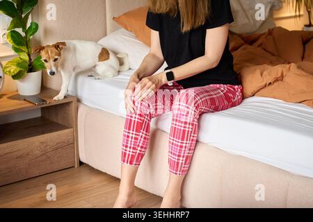 La donna soffre di dolore al ginocchio mentre si siede a letto vicino a un cane piccolo, tenendo il ginocchio in camera da letto. Concetto di disagio articolare, dolore alle gambe e assistenza sanitaria. Foto Stock